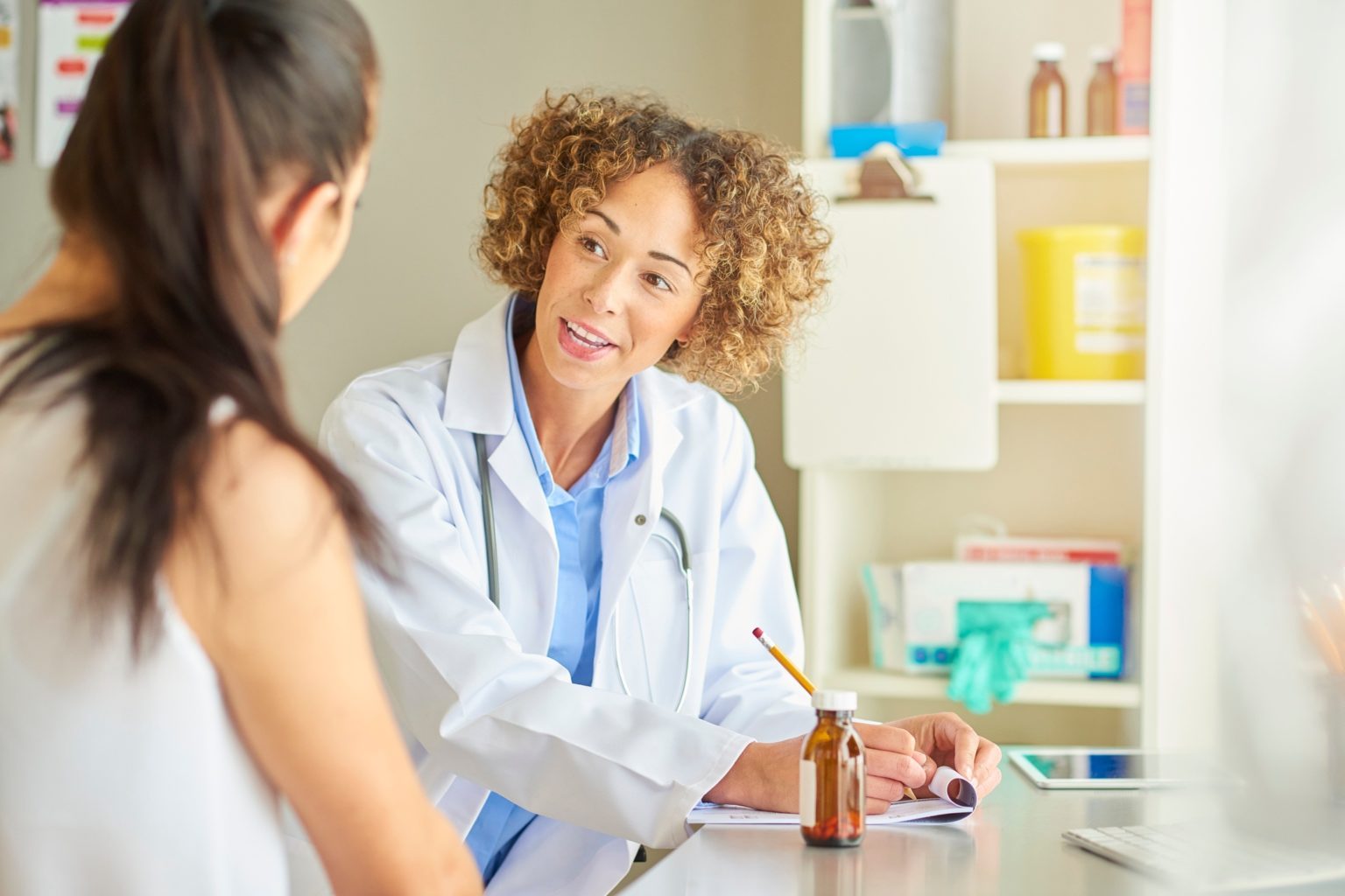 Doctor in a white coat leaning in and speaking with a patient across an exam room desk while taking notes