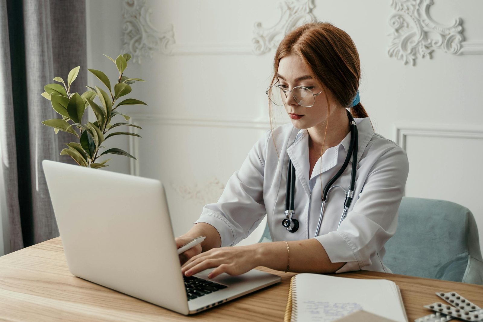 Physician in a white coat and stethoscope typing on a laptop at a desk, a notebook and blister packs beside her