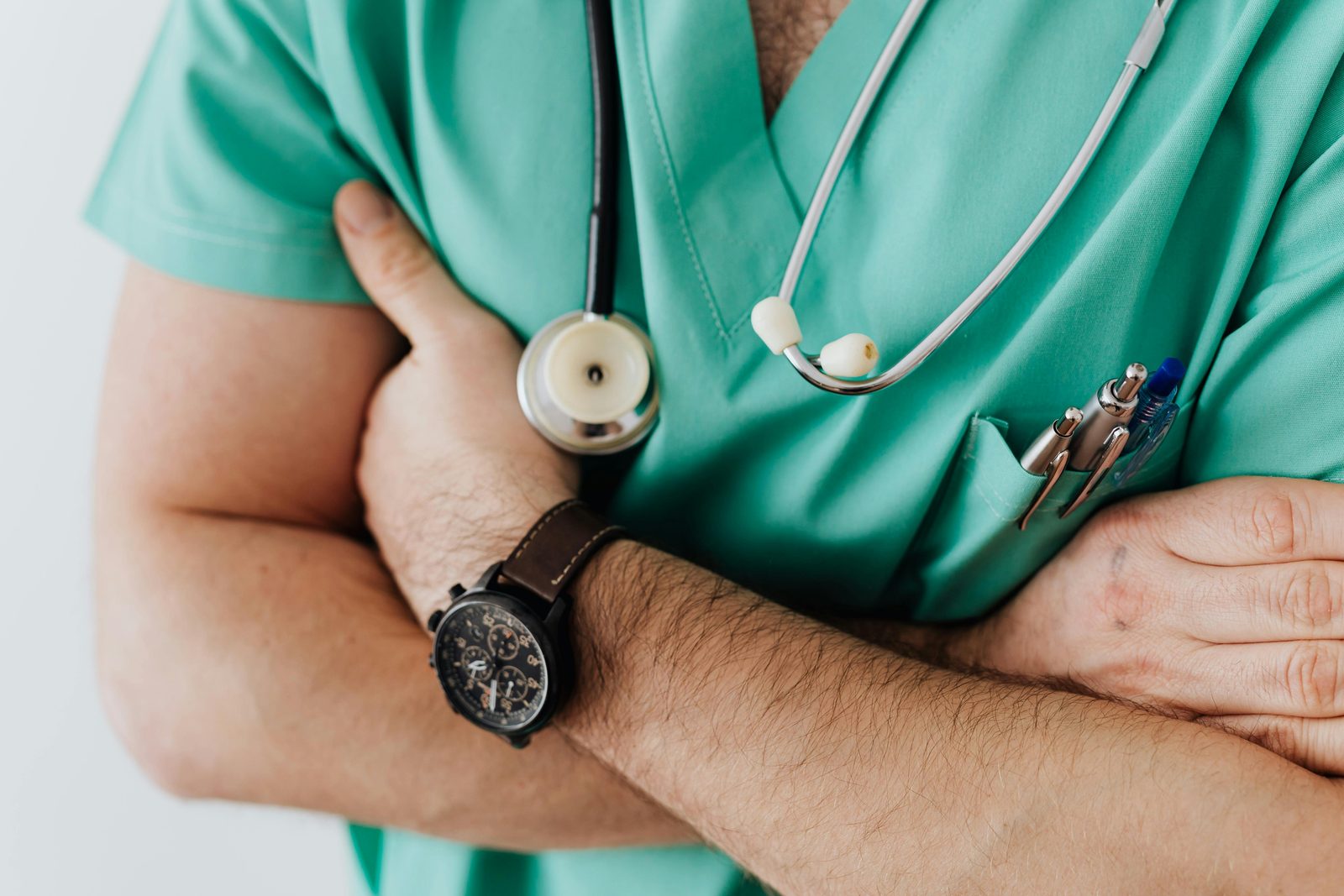 Close-up of a clinician in green scrubs standing with arms folded, a stethoscope around the neck and a watch on the wrist.