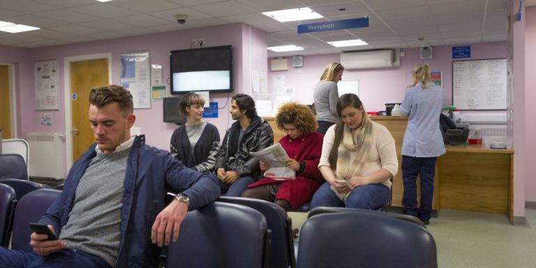 Patients seated in a clinic waiting room, most looking at their phones or a magazine, with staff at the reception desk behind them.