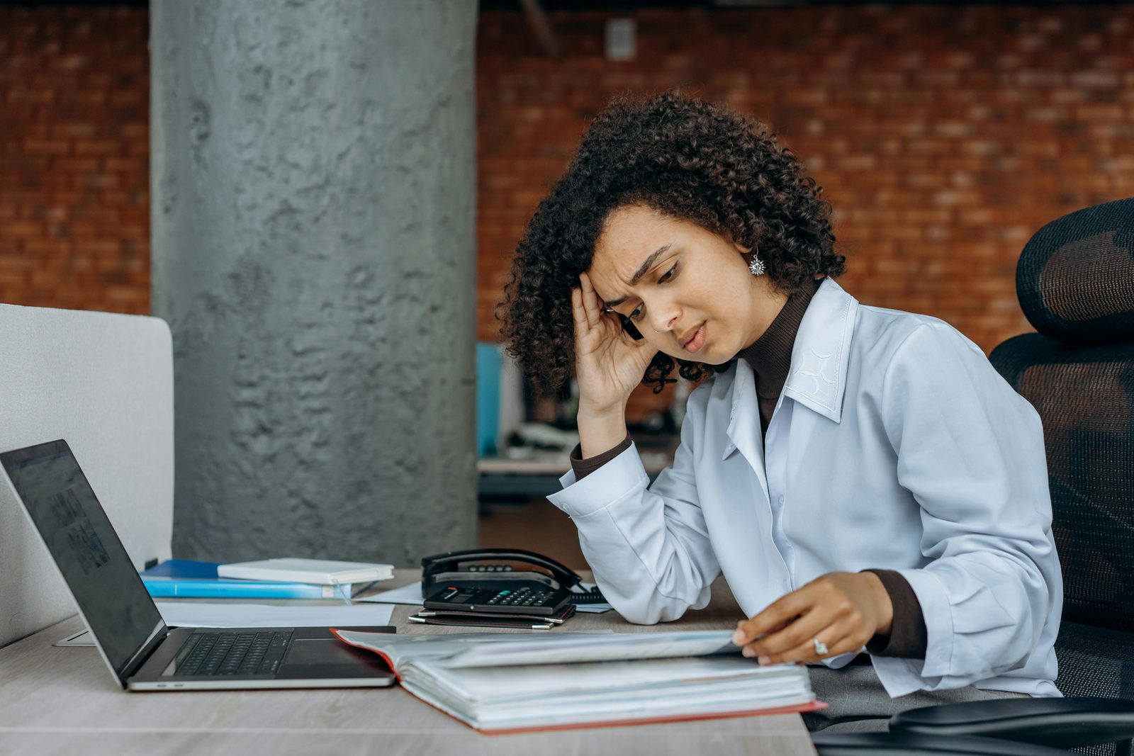 Clinician resting her head on one hand at a desk, paging through a thick binder of paperwork beside a laptop